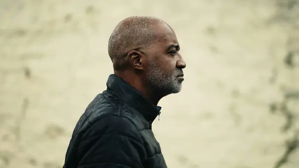 One pensive middle-aged black man walking forward in urban street with thoughtful gaze. Tracking shot close-up face of a senior mature person of African descent strolling outside