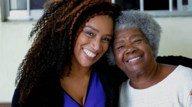 80 'lerin yaşlı, kır saçlı annesinin kollarına sarılmış yetişkin Afro-Amerikan kızının mutlu portresi. Kolları omzunda neşeli nesiller arası kadınların yakın çekim yüzleri.