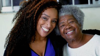 80 'lerin yaşlı, kır saçlı annesinin kollarına sarılmış yetişkin Afro-Amerikan kızının mutlu portresi. Kolları omzunda neşeli nesiller arası kadınların yakın çekim yüzleri.