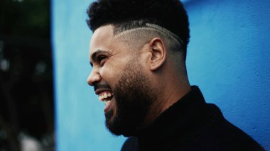 Mutlu bir Latin asıllı genç adam kamerasız biriyle konuşurken gülüyor ve gülümsüyor.