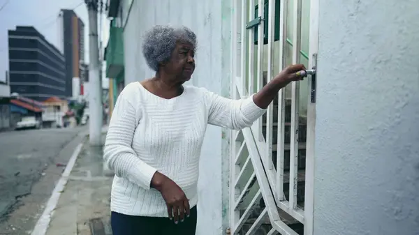 One elderly black lady opening residence front door, arriving home from urban sidewalk street. 80s African American woman returns to South American house after daily routine activity