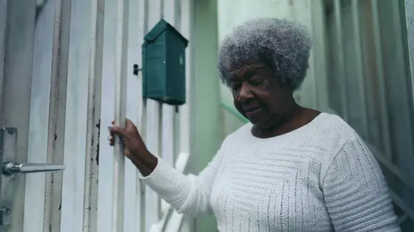 One elderly black woman from South America steps out in the street from residence front door into urban sidewalk, senior lady going for her daily active walk