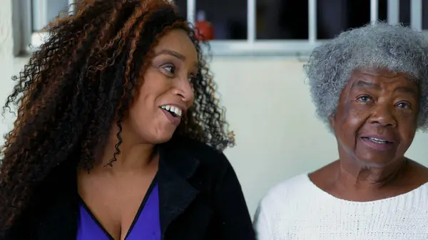African American Adult daughter authentic interaction with elderly 80s mother exchanging glances while laughing and smiling, genuine happy authentic faces of contrasting ages