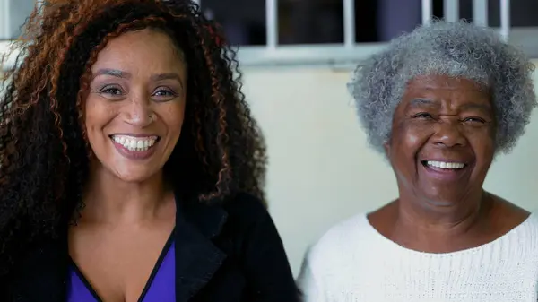 African American Adult daughter authentic interaction with elderly 80s mother exchanging glances while laughing and smiling, genuine happy authentic faces of contrasting ages