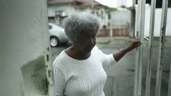 One elderly black lady opening residence front door from urban sidewalk and climbs up the stairs returning home. Humble Senior gray-hair African American woman arrives at residence