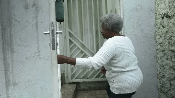 Elderly South American black woman arrives home from sidewalk street, opens residence front door returning by closing gate behind her, senior 80s person of African descent