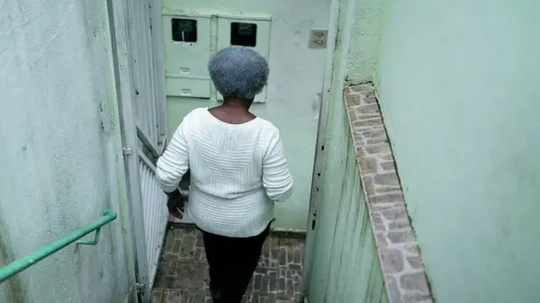 One elderly black lady stepping out to the street from residence. Back of gray-hair African American woman opening front door and going for a walk outside in street