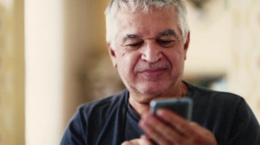 Kafkasyalı kıdemli bir adam, modern teknolojiye dalmış, yüzüne yakın çekim yaparken neşeli bir sırıtış sergiliyor, evinde cep telefonuna bakıyor.