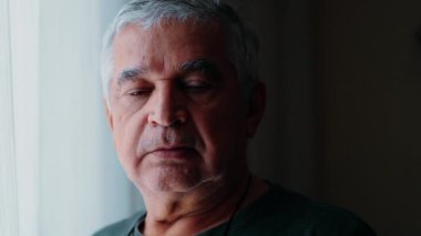 Yaşlı gri saçlı adam perdenin yanında duran kameraya bakıyor. Pencere resmi yakın plan, kırışık 70 'li yaşlarda.