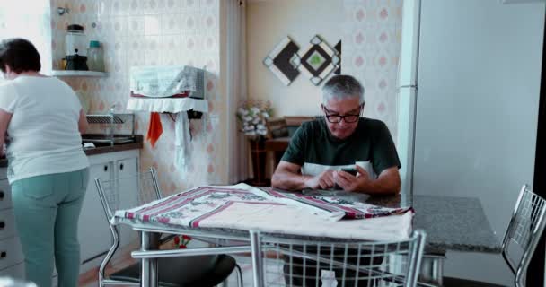 Hombre Mayor Sentado Cocina Mirando Dispositivo Móvil Pintoresca Escena Residencia — Vídeos de Stock