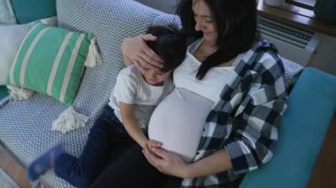 Evdeki kanepede oturan doğmamış kardeşime sevgi göstergesi olarak hamile bir anneye sarılan şefkatli bir çocuk.
