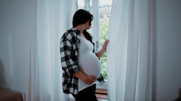 ウィンドウカーテンで立っている妊娠中の女性は 家の外を眺めている 産婦人シーン ヶ月腹 — ストック動画