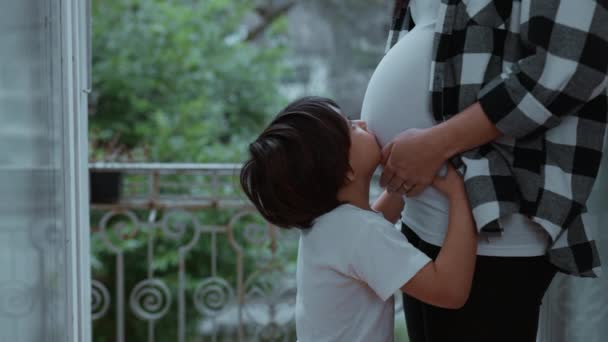 赤ちゃんへの愛と愛情のディスプレイで母親の妊娠中の腹にキス小さな男の子 妊娠の第三期の間に ママと家庭のバルコニーに立って — ストック動画