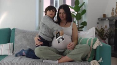Mutlu anne ve çocuk, hamileliğin üçüncü üç ayı boyunca evde oturan doğmamış erkek kardeşin ultrason fotoğrafıyla poz veriyor. Anne ve oğlu kameraya gülümsüyor.