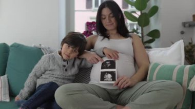 Ultrason Fotoğraflı Mutlu Anne ve Çocuk Pozisyonu - Doğmamış Bebek Kardeşimin İmajıyla Kameraya Gülümseme, Üçüncü Trimestr 'de Ev Kanepesinde Oturma