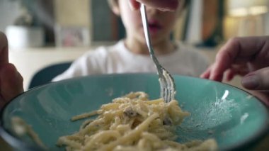 Mavi tabakta makarnayı çeviren çatallı spagettinin yakın çekimi bulanık arka planda küçük bir çocuk, annenin eli oğluna makarna çevirmeyi öğretiyor, çocuk yemeği zamanı.