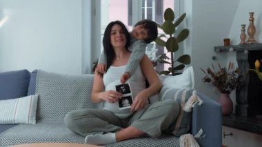 Anne ve çocuk kameraya poz veriyor. Üçüncü üç aylık hamilelik döneminde ultrason çekiliyor. 30 'lu yaşlardaki anne ve 5 yaşındaki oğlu, bebek kardeşi için kanepeye oturuyor.