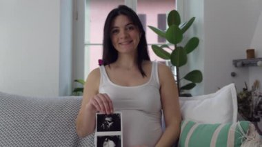 Yakında anne olacak kişi, Ultrason görüntüsü sunacak 30 'lu yaşlardaki anne karnını okşayacak ve doğmamış bebeğe bakacak, Evdeki Kanepede mutlu bir şekilde oturacak.