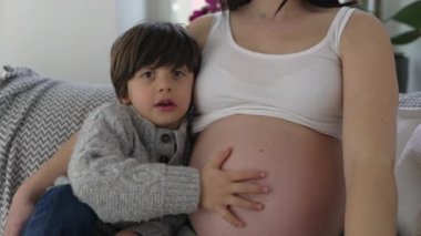 Küçük çocuk annesinin hamile karnına hafifçe dokunuyor. Hamileliğinin sonlarına doğru doğmamış erkek kardeşine sevgi ve ilgi gösteriyor.