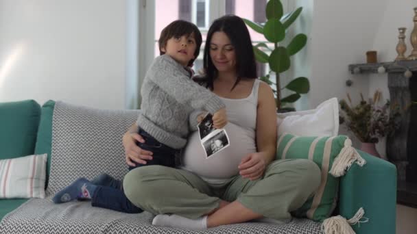 ハッピーマザーと子供は 自宅のソファに座っている妊娠の第3期の間に生まれていない兄弟の超音波写真をポーズします カメラで笑顔のママと息子 — ストック動画