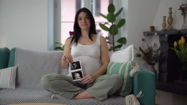 임신의 삼분기 집에서 소파에 앉아있는 아기의 초음파 사진을 보여주는 행복한 — 비디오