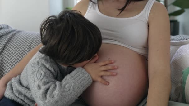 5岁男孩在怀孕后期轻吻妈妈的腹部 孩子和妈妈坐在沙发上等未出生的弟弟 母爱的概念 — 图库视频影像
