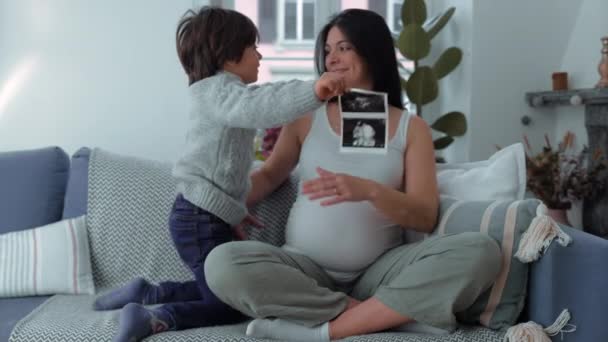 超音波写真を持つ母と子供 30代のママと5歳の息子 期待する赤ちゃんの兄弟 サードトリムスターのホームカウチにポジショニング — ストック動画