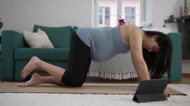 Hamile kadın evde egzersiz yapıyor, doğum öncesi antrenman sırasında bacağını kaldırıyor. Evde, oturma odasının zemininde, yoga minderinde tablet aletinin önünde.