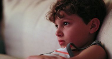 Child watching TV screen, casual candid young boy seated at living-room sofa