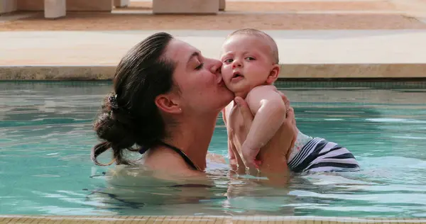Mother kissing newborn baby son inside swimming pool water