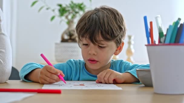 小さな男の子はカラフルなマーカーのバケツの横のテーブルで着色する宿題を楽しんでいます — ストック動画