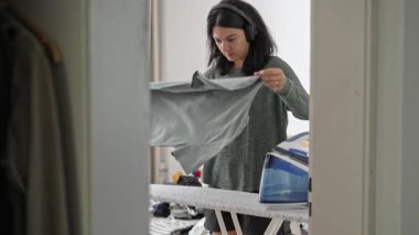 Kulaklık takan bir kadın yeni ütülenmiş gömleğini alıyor ve dükkanda katlıyor.