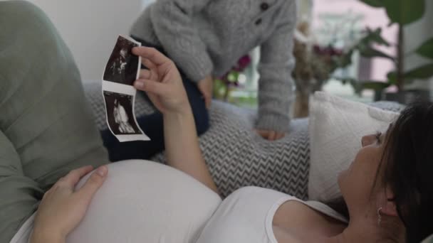 母と息子は 妊娠後期にソファーで休憩しながら 赤ちゃんの兄弟の超音波を見ながら幸せな柔らかい瞬間を共有しています 穏やかに腹に触れる子供 — ストック動画