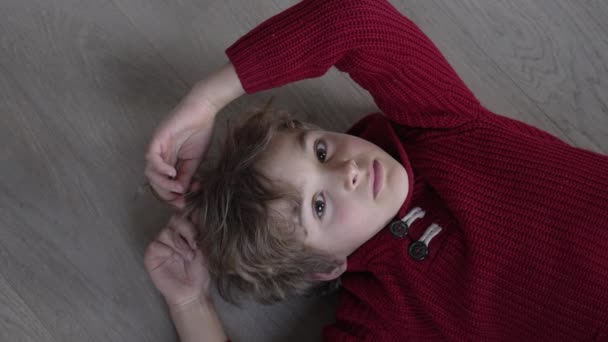 一个多愁善感的小男孩一边玩着头发 一边看着放在地板上的摄像机 穿着红色毛衣 表情体贴的孩子 做白日梦的孩子 — 图库视频影像