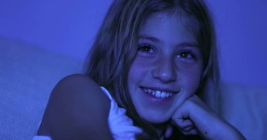 Küçük kız, karanlıkta televizyon izlerken gülüyor. Gerçek hayatta ve samimi bir gülümsemede.