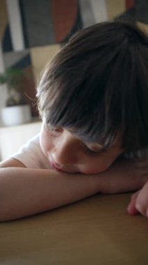 Üzgün küçük bir çocuk aldatma ifadesiyle masaya yaslanmış, sıkılmış 5 yaşındaki üzgün çocuk, dikey video