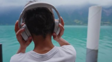 Kulaklıklardan kulaklıkları çıkaran, güzel manzaralı göl manzarasına bakan, sese ara veren ve doğayı takdir eden genç bir çocuk.