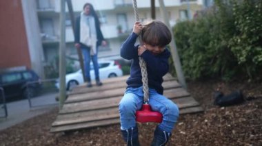 Kaygısız küçük çocuk, sonbahar günü kamu parkında iki ağacın arasına bağlanmış halatı sıkıca tutarak tel ipten aşağı kayıyor. Çocuk eğleniyor.