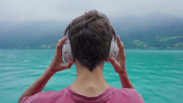 年轻男子站在湖边看风景时 背着耳机捂住耳朵 在风景秀丽的自然景观中听音乐 有声读物或播客的人 — 图库视频影像