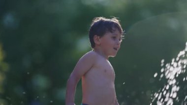 Küçük çocuk, yavaş çekim 120 fps yüzme havuzu çeşmesinde kendini yeniliyor. Sıcak yaz gününde su damlacıklarının keyfini çıkaran bir çocuk.