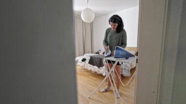 Konforlu ev elbiseli bir bayan, kulaklıkla bir şeyler dinlerken odasında sakince ütülüyor.