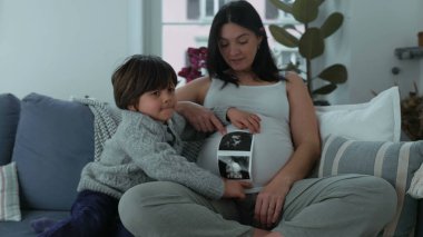 Evdeki koltukta oturan hamile bir annenin yanında oturan bir çocuğun ultrason görüntüsü. Gerçek bir aile bağının doğmamış erkek kardeşi beklediği an.