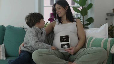 Hamileliğin üçüncü üç ayında hamile bir anneye sarılan 5 yaşındaki oğlu, evdeki koltukta oturan çocuk ve anne için poz veren annesi, ultrason fotoğrafı tutan annesi.
