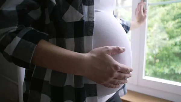 stock image Close-up of mother caressing belly, expecting newborn child - 8 month pregnancy by residence window