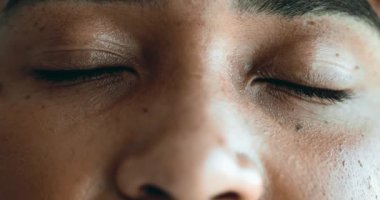 Meditasyon yapan genç siyah bir kadın rahatlama ve tefekkür için gözlerini kapatıyor. Makro yakın plan yüz hatları sessizce dinlenirken, kameraya gülümseyerek gözlerini açar.