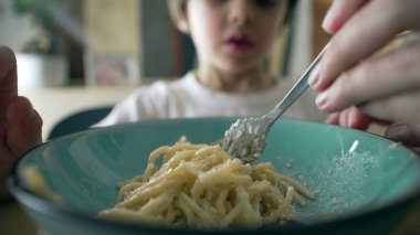 Mavi tabakta makarnayı çeviren çatallı spagettinin yakın çekimi bulanık arka planda küçük bir çocuk, annenin eli oğluna makarna çevirmeyi öğretiyor, çocuk yemeği zamanı.