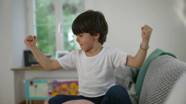 Sevimli 5 yaşında küçük bir çocuk kollarını sallayarak evde gücünü gösteriyor ve kaslarının gururlu bir şekilde büyüdüğünü gösteriyor.