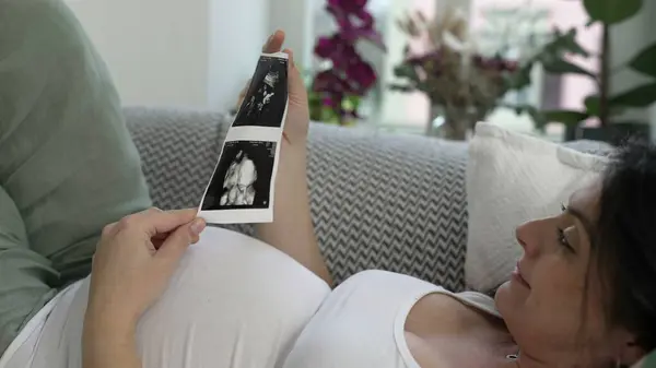 stock image Pregnant woman looks at ultrasound image of her baby during late stage pregnancy laid on couch resting at home embracing maternal joy