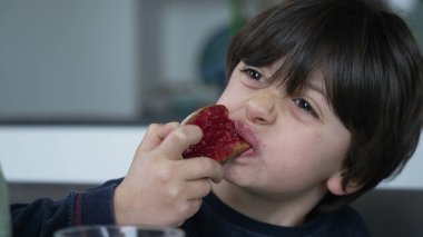 Kahvaltı masasında reçelli tost yiyen küçük bir çocuk, çocuğun yakın plan yüzü bir lokma yiyecek.