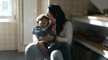 Anne, 5 yaşındaki oğlunu mutfakta tutarken dinleniyor. Otantik bir aile hayatı sürüyor. Yorgun bir anne, gözleri kapalı ve çocuğu alnından öpüyor.